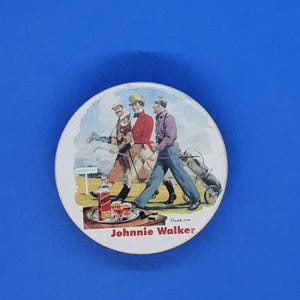 May include: Round coaster featuring a vintage illustration of three golfers walking on a green golf course. A tray with Johnnie Walker whiskey and glasses is in the foreground. The text "Johnnie Walker" is at the bottom.