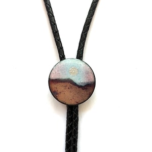 May include: A bolo tie featuring a circular pendant with a landscape design. The pendant displays a textured brown lower section and a light blue upper section, separated by a dark line. A small silver circular accent sits in the blue area. The cord is black braided leather.