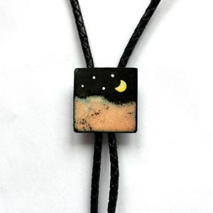 May include: A bolo tie featuring a square pendant with a night sky design. The pendant has a black background with white stars and a yellow crescent moon. The bottom portion is tan. The tie has a black braided leather cord.