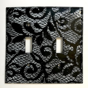 May include: A decorative double light switch cover with a black and white lace pattern. The cover has two switch openings and screw holes for mounting. The design features a detailed, swirling lace motif.