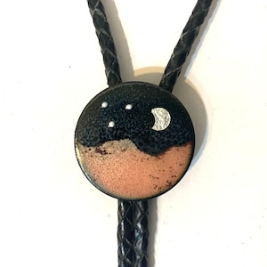 May include: A round bolo tie pendant with a night sky design. The top half is dark blue with three white dots and a silver crescent moon. The bottom half is a copper color. The braided black leather cord is visible.
