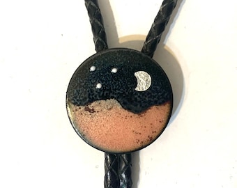 Op de afbeelding: Ronde bolo stropdas hanger met een nachthemel ontwerp. De bovenste helft is donkerblauw met drie witte stippen en een zilveren halve maan. De onderste helft is koperkleurig. Het gevlochten zwarte leren koord is zichtbaar.