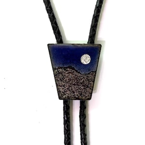 May include: A bolo tie with a geometric pendant. The pendant features a dark blue upper section and a textured, speckled lower section. A silver-colored circle is on the blue section. The tie has a black braided cord.