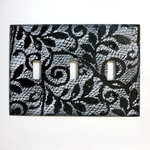 May include: A decorative triple light switch cover with a black and white lace pattern. The design features a detailed floral and scrollwork motif, with three rectangular switch openings and mounting holes.