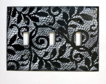 May include: A decorative triple light switch cover with a black and white lace pattern. The design features a detailed floral and scrollwork motif, with three rectangular switch openings and mounting holes.