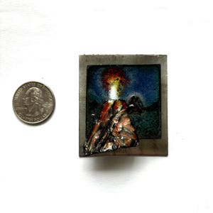 May include: A small metal framed artwork depicting a volcano erupting with red, orange, and yellow lava against a blue and green background.