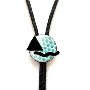 May include: A black leather bolo tie with a circular pendant. The pendant features a white background with teal polka dots, a black triangle, and a black curved line. The bolo tie is a unique accessory.