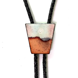 May include: A bolo tie featuring a trapezoidal pendant with an enameled surface displaying a landscape of white, pale pink, and terracotta orange tones with a small circular detail, suspended from a black braided leather cord.