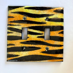 May include: A decorative double light switch cover with a tiger stripe design. The cover features a glossy finish with alternating black and yellow stripes. Includes two switch openings and four screw holes for mounting.