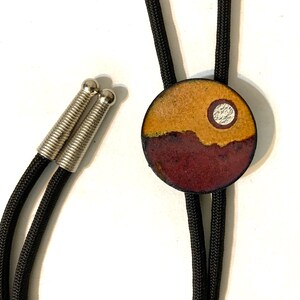 May include: A black bolo tie with a round ceramic pendant featuring a landscape scene with a yellow sky, brown hills, and a white circle.