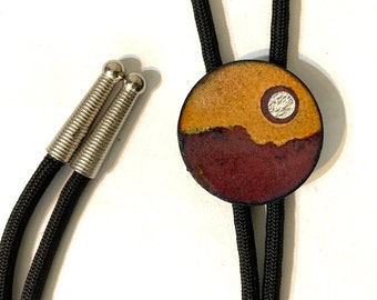 May include: A bolo tie featuring a circular pendant with a two-tone enamel finish in mustard yellow and deep maroon, accented by a small silver-toned circle. The pendant is attached to a black braided cord with silver-toned metal tips at the ends.