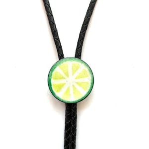 May include: A bolo tie featuring a circular pendant with a lime slice design in shades of light green, yellow, and white. The pendant is attached to a black braided leather cord that extends upwards and downwards from the center piece.