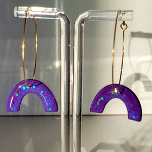 Könnte beinhalten: Ein Paar violette Harzhüpfringe mit goldenen Ringen und schimmerndem Glitzer. Die Ohrringe sind halbkreisförmig.