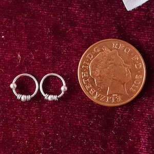 Puede incluir: Dos pequeños pendientes de aro plateados con un diseño de cuentas se muestran sobre una superficie de terciopelo rojo oscuro. También hay una moneda de cobre y acentos de flores blancas. Los pendientes son delicados y parecen hechos a mano.