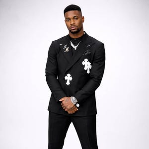May include: Black double-breasted suit with white cross and initial embellishments. The suit features a notched lapel, a pocket square, and a silver watch. The suit is paired with a black shirt and a silver chain necklace. The overall look is stylish and modern.