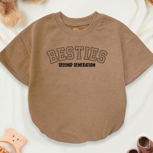 May include: A tan baby romper with the word "BESTIES" in black block letters and the words "SECOND GENERATION" underneath. The romper is surrounded by a teddy bear, a wooden toy, and a pair of brown baby shoes.