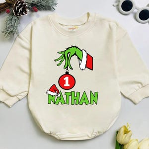 May include: Cream-colored baby romper with a Grinch-themed design. Features a green hand holding a red and white ornament with the number 1. The name "Nathan" is printed in green below. A small red Santa hat is also present.