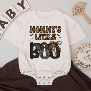 May include: White baby onesie with the text "Mommy's Little Boo" in orange and black. The word "Boo" features bat wings and a bow. Wooden blocks spell "BABY" and a wooden rattle are in the background. A Halloween-themed baby outfit.