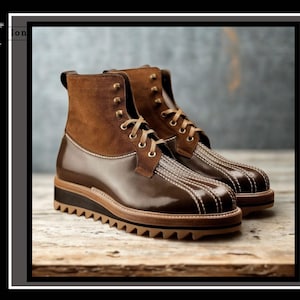 May include: A pair of ankle-high boots featuring a dark brown polished leather lower section with prominent white stitching and a brown suede upper. The boots include brown laces, brass-toned eyelets, and a thick, tan-colored serrated rubber sole.