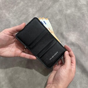 Può includere: Portafoglio nero con la scritta "PHENOM" stampata nell'angolo in basso a destra. Il portafoglio è aperto, rivelando fessure per carte e una tasca. Diverse banconote in euro sono visibili all'interno. Il portafoglio è tenuto aperto da due mani.