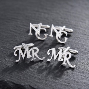 May include: Three pairs of silver cufflinks are displayed on a dark gray surface. Two pairs feature the initials "NG", and the third pair displays "MR". The cufflinks have a polished finish and a classic design.