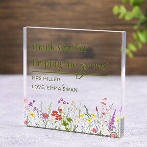 May include: Clear acrylic block with the text "thank you for helping me grow" in green. The block is personalized with "MRS MILLER" and "LOVE, EMMA SWAN". A colorful floral design decorates the bottom edge. The block is a thoughtful gift.