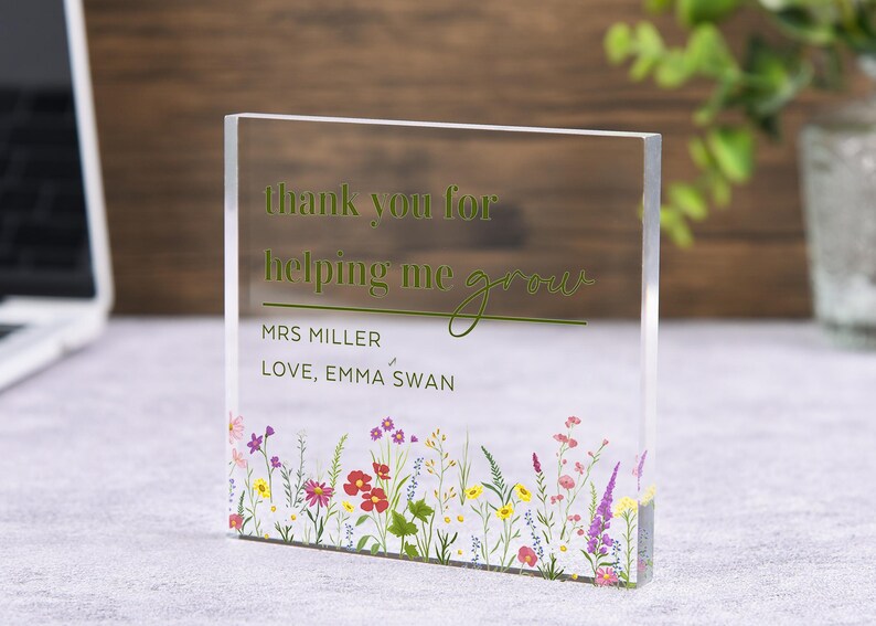 May include: Clear acrylic block with the text "thank you for helping me grow" in green. The block is personalized with "MRS MILLER" and "LOVE, EMMA SWAN". A colorful floral design decorates the bottom edge. The block is a thoughtful gift.