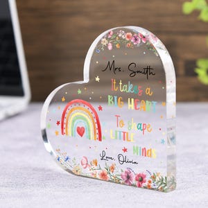 May include: A heart-shaped acrylic block with a colorful rainbow design and floral accents. The block features the text "Mrs. Smith, It takes a BIG HEART To shape LITTLE Minds, Love, Olivia." A thoughtful gift for a teacher.