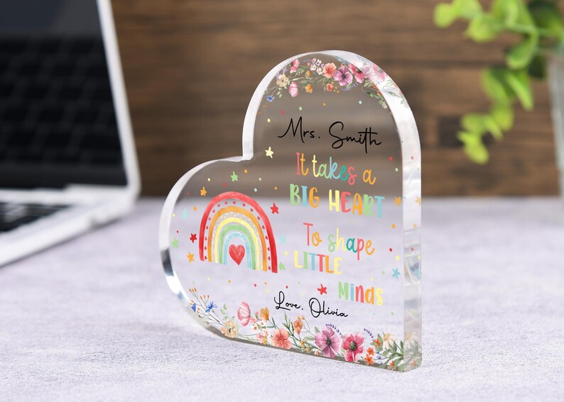 May include: A heart-shaped acrylic block with a colorful rainbow design and floral accents. The block features the text "Mrs. Smith, It takes a BIG HEART To shape LITTLE Minds, Love, Olivia." A thoughtful gift for a teacher.