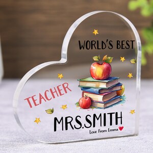 May include: Clear acrylic heart-shaped plaque with the words "WORLD'S BEST TEACHER" and "MRS. SMITH" in black. A stack of books and apples are illustrated on the plaque. The text "Love From Emma" is also included.