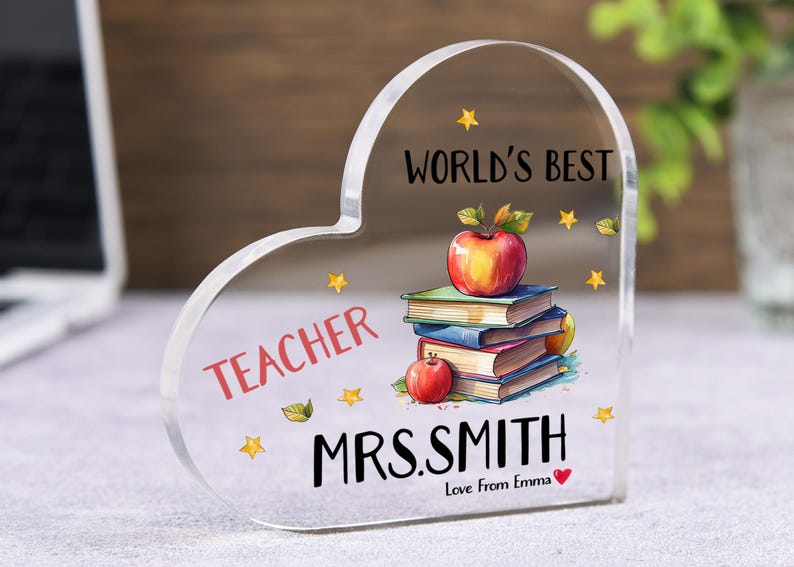 May include: Clear acrylic heart-shaped plaque with the words "WORLD'S BEST TEACHER" and "MRS. SMITH" in black. A stack of books and apples are illustrated on the plaque. The text "Love From Emma" is also included.