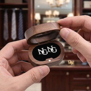 May include: A wooden jewelry box, oval-shaped, with a hinged lid. The interior is lined with black velvet and features the letters "N&NG" in a white, stylized font. The box is held open, revealing the contents.