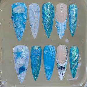May include: A set of ten long, pointed acrylic nails with ocean-themed designs. The nails feature shades of blue, white, and clear, with embellishments like glitter, rhinestones, and sculpted wave and sea creature details.