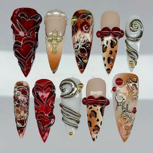 May include: A set of ten long, pointed artificial nails with various designs. The designs include red hearts, gold crosses, leopard print, and swirling silver and gold accents. Some nails feature pearl and bow details.