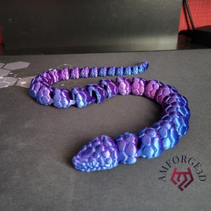 May include: A 3D-printed articulated snake toy in shades of blue and purple. The snake's body is segmented with a honeycomb pattern, and it is lying on a dark surface. The logo "AMFORGE3D" is visible.