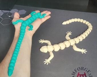 May include: Two articulated lizard toys. One is teal and held in a hand, the other is off-white and lying flat. Both have segmented bodies and poseable limbs. The logo "AMFORGE3D" is visible in the bottom right corner.