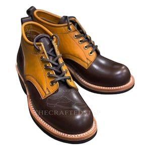 May include: A pair of two-tone leather boots. The boots feature a brown toe and heel, with a tan-colored upper. The boots have dark gray laces and are stitched with a light brown thread. The soles are a light brown color.