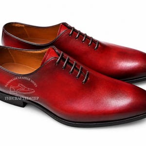 May include: A pair of red leather dress shoes with dark laces. The shoes have a polished finish and a classic Oxford design. The text "HANDMADE LEATHER SHOES" and "THECRAFTEDSTEP" is visible on the side of the shoe.