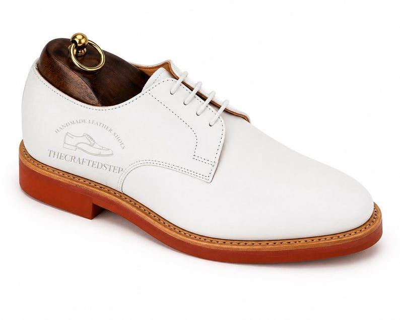 May include: White leather derby shoe with a brown sole and a wooden shoe tree. The shoe has white laces and the text "HANDMADE LEATHER SHOES" and "THE CRAFTED STEP" printed on the side. The shoe tree has a gold-colored ring.