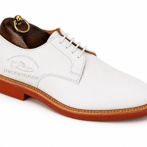 May include: White leather derby shoe with a brown sole and a wooden shoe tree. The shoe has white laces and the text "HANDMADE LEATHER SHOES" and "THE CRAFTED STEP" printed on the side. The shoe tree has a gold-colored ring.