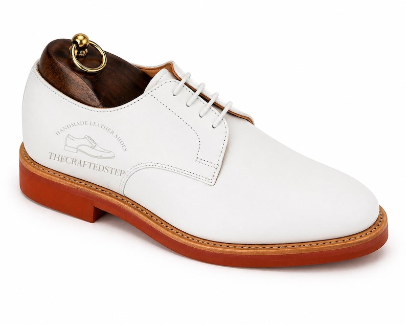 May include: White leather derby shoe with a brown sole and a wooden shoe tree. The shoe has white laces and the text "HANDMADE LEATHER SHOES" and "THE CRAFTED STEP" printed on the side. The shoe tree has a gold-colored ring.