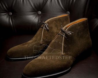 May include: A pair of brown suede chukka boots with black laces and soles. The boots have a classic design and are made of a soft, textured material. The text "HANDMADE LEATHER SHOES" and "THE CRAFTEDSTEP" is visible.