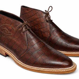 Könnte beinhalten: Braune Leder-Chukka-Stiefel mit einem strukturierten, krokodilartigen Muster. Die Stiefel haben eine hellfarbige Sohle und Schnürsenkel. Der Text "HANDMADE LEATHER SHOES" und "THE CRAFTED STEP" ist auf der Seite eingeprägt.