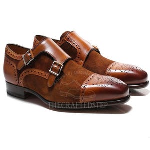 Peut inclure: Une paire de chaussures marron en cuir à double boucle avec des empiècements en daim. Les chaussures présentent un bout de capuchon avec des perforations décoratives et des boucles argentées. Les chaussures sont présentées sur fond blanc.