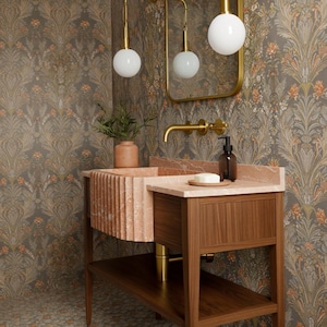 May include: A bathroom vanity with a pink marble sink and countertop, set against a floral wallpaper. The vanity has a wooden base with a lower shelf. A gold faucet and light fixtures complement the design. A soap dispenser and a small vase are on the countertop.