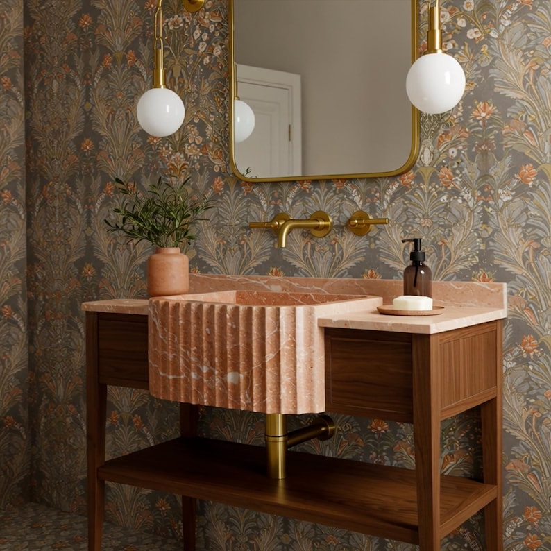May include: A bathroom vanity with a pink marble sink and countertop, set against a floral wallpaper. The vanity has a wooden base with a lower shelf. A gold-framed mirror and matching fixtures complete the elegant design.