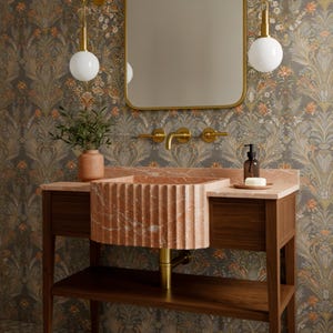 May include: A bathroom vanity with a fluted, reddish-orange marble sink. The sink sits on a wooden cabinet with a lower shelf. A gold-framed mirror and matching faucet fixtures are mounted on the wall. The wallpaper has a floral pattern.