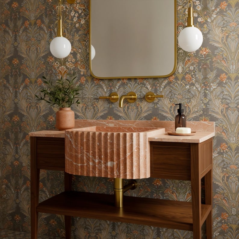 May include: A bathroom vanity with a fluted, reddish-orange marble sink. The sink sits on a wooden cabinet with a lower shelf. A gold-framed mirror and matching faucet fixtures are mounted on the wall. The wallpaper has a floral pattern.