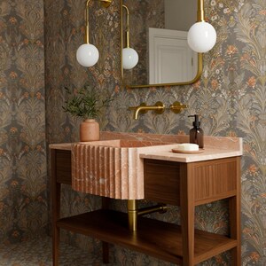 May include: A bathroom vanity with a pink marble sink and countertop, set against a floral wallpaper. The vanity has a wooden base with a lower shelf and gold fixtures, including a faucet and pendant lights.