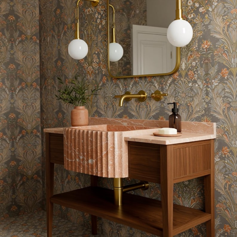 May include: A bathroom vanity with a pink marble sink and countertop, set against a floral wallpaper. The vanity has a wooden base with a lower shelf and gold fixtures, including a faucet and pendant lights.